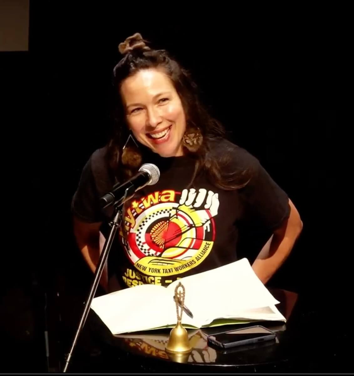 Emily Johnson stands at a podium and smiles as she speaks into a microphone on a stand. She has light skin and long brown hair which is partially pulled back, and she is wearing a black short-sleeved t-shirt bearing the logo of the New York Taxi Workers Alliance. Before her on the podium is an open book, her phone, and a gold colored long-handled bell.