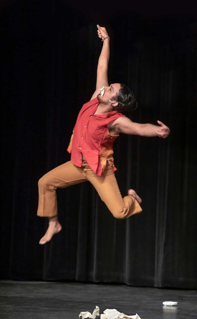 A medium, brown-skinned Mexican American dancer leaps looking upwards with a mouth full of torn pages of a book. His gaze is looking left with his right arm reaching upholding a fist and the other extending out from shoulder height. His legs are bent in a kneeling position suspended from the ground. He wears a short sleeve red button-up top with a single wide vertical orange stripe along the rib cage his pants are wide-leg and orange. The background has a black curtain and a dark floor with torn pages at the edge.