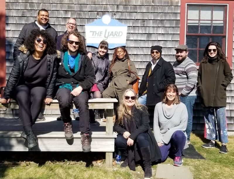 A diverse group of eleven adults pose for a photo on and around an unpainted picnic table  in front of building clad in weathered wooden shingles with red wooden framed square windows. There is a blue and white sign on the wall that says "The Yard." They are all dressed snugly but not in a way that implies it's very cold outside. It is quite bright, though, and several of them are wearing sunglasses. They are all smiling or laughing.