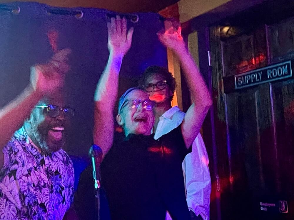 In a partially blurred photo that looks like it was taken in a dark room without a flash, a black man and a white man stand against a makeshift backdrop and raise their hands as they sing together into a microphone. They are smiling as they sing. Both of them are wearing glasses. The black man on the left is wearing a short sleeved, buttoned shirt covered in an intricate black and white design of plant leaves. The white man on the right is wearing a short-sleeved black shirt. Behind them, a tall black woman wearing a loose-fitting, white, long-sleeved shirt and glasses watches them and smiles. To her left--on the right side of the image--is a wooden door marked "Supply Room."