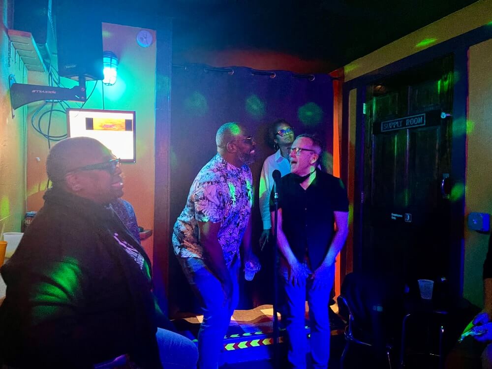 In a dimly lit photo that looks like it was taken in a darkened room, a black man and a white man stand against a makeshift backdrop and bend slightly with their hands placed just above their knees and look at each other, smiling, as they sing together into a microphone. Both of them are wearing glasses. The black man on the left is wearing a short sleeved, buttoned shirt covered in an intricate black and white design of plant leaves. The white man on the right is wearing a short-sleeved black shirt. To the left of them, in the foreground, a black woman with a shaved head, black glasses, and a shiny, leather-like black jacket smiles in profile as she looks up at what seems to be a karaoke monitor. Behind the men, a tall black woman wearing a loose-fitting, white, long-sleeved shirt and glasses stares over their heads and to the left as if reading the lyrics from an unseen karaoke monitor. To her left--on the right side of the image--is a wooden door marked "Supply Room."