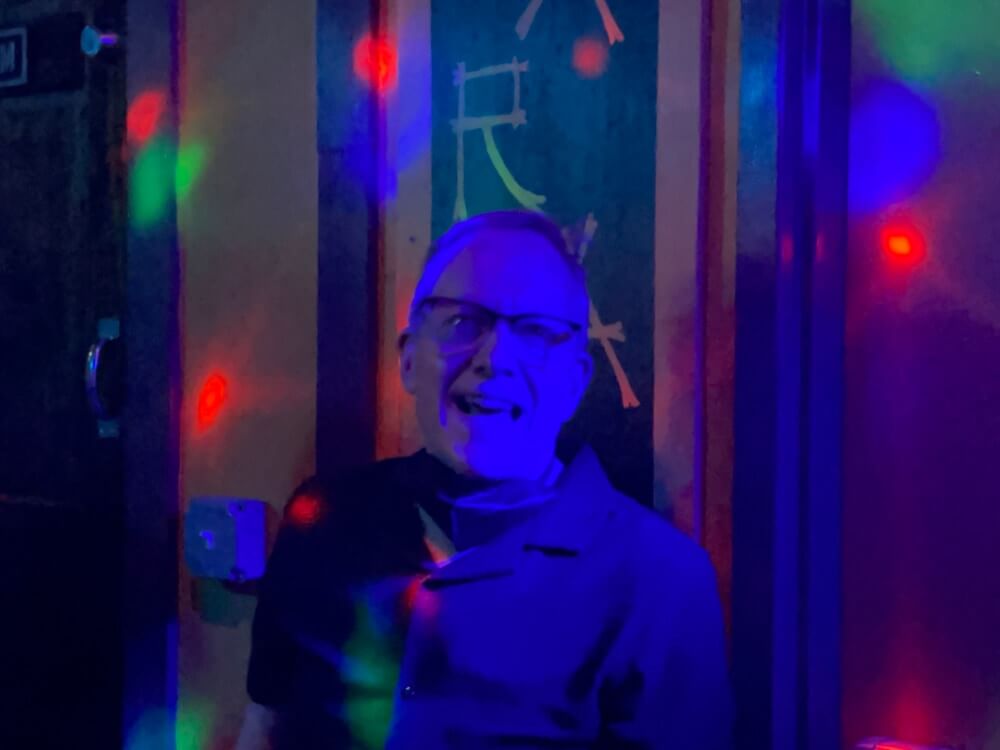 A white man with short hair and horn-rimmed half-frame glasses smiles broadly at the camera, as if caught mid-laughter or in the middle of saying something. There are spots of colored light on the wall behind him, suggesting the environment is a bar or club. Due to the unusual lighting, all the highlights of the image are replaced with a deep blue color, and the blacks are a dark bluish-gray, which gives the photo a heavily stylized, almost painterly effect.
