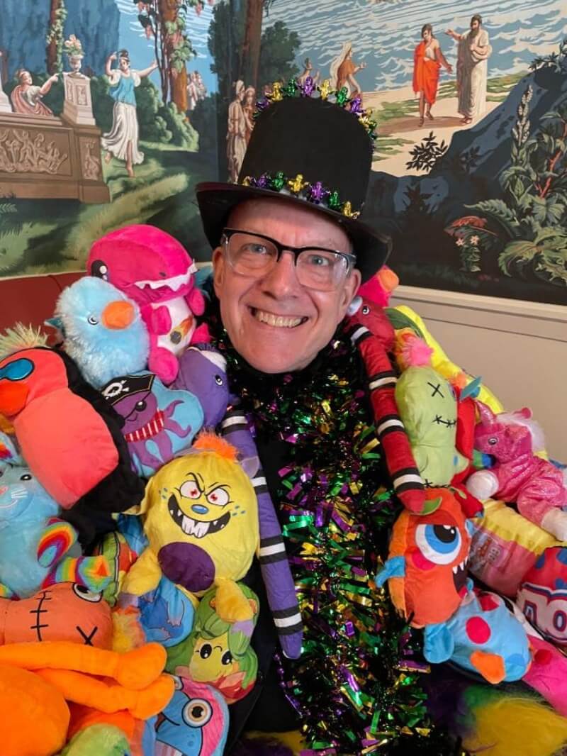 A medium close-up of a middle-aged, white man with horn-rimmed, half-frame glasses smiles for the camera. He is dressed in an elaborate Mardi Gras costume, and based on the angle of his body and how close he is to the corner of the room, it appears that he is sitting for this photo. His bulky but soft jacket is covered entirely with stuffed animals in every color, and it's open to reveal a shirt or wide scarf made of purple, green, and yellow metallic tinsel. He is wearing a black felt top hat decorated with a band of metallic purple, green, and yellow beads. Behind him on the upper half of the adjoining walls is a mural of what looks like a Greek pastoral scene rendered in a slightly desaturated illustration style. 