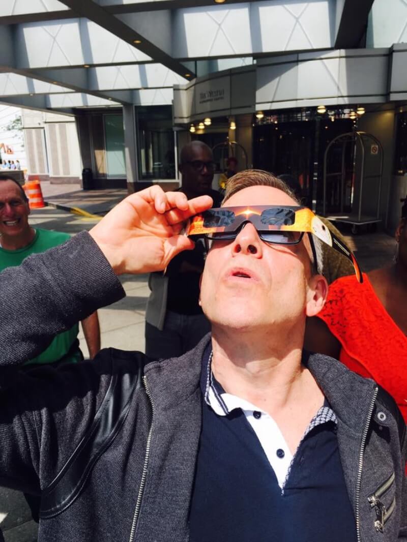 A white man with short, light-colored skin  and black glasses is standing outside under or right next to the awning of a hotel. Behind him in the shadows are other people looking off at something or staring at him and smiling. He is holding a pair of disposable eclipse glasses over his own glasses and looking up at the sky with a look of focused effort. He wears a blue polo shirt with a white placket, and over that he is wearing an opened, dark gray, long-sleeved jacket with a zipper front.