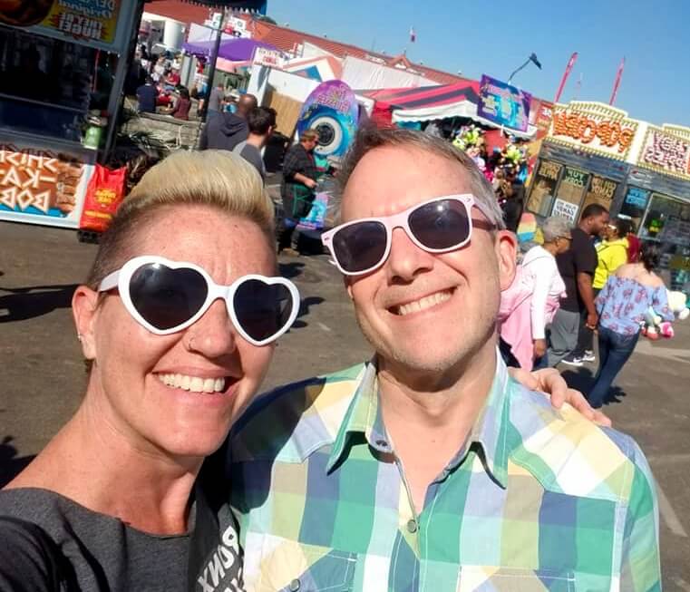 A smiling white woman wearing white heart-shaped sunglasses poses for a selfie with a white man who is also smiling and wearing light pink sunglasses. The woman is on the left, and her hair is two-tone: on the sides it is dark and trimmed close to the scalp, and on top it is blonde and longer, but swept back in a low pompadour. She is wearing a collarless dark gray t-shirt with indecipherable white lettering on the front. The man has short hair that's swept up off his forehead, and he is wearing a button down green and yellow plaid shirt. They are standing in what appears to be the midway at a small carnival, and behind them in the middle distance are a shish-ka-bob stand on the left and a popcorn stand on the right. Between them and the food stands there are two small groups of people walking by.