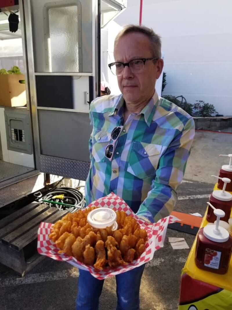 A medium close-up of a white, middle-aged man with short, lightly colored hair, and black, horn-rimmed, half-frame glasses, is outside, walking past the camera and away from a condiment stand with large plastic pumps of ketchup on his left, and the open door of a food truck on his right. He is wearing a green, blue, and yellow plaid, long-sleeve, button-down shirt, and a pair of light pink sunglasses are hooked over the open collar. He is holding a large platter covered in red and white checked paper, on top of which has been arranged in a circular pattern dozens of pieces of identically shaped, unidentified fried food, with plastic containers of dipping sauce in the center. He appears to be concentrating on carrying the dish of food. 