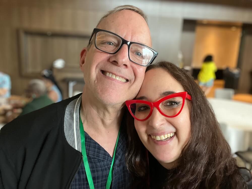 A smiling, white man on the left side of the image poses for a selfie with a slightly darker skinned smiling woman on the right. The man is wearing black framed glasses and has a green lanyard around his neck, and he's wearing a dark blue and black tightly-patterned plaid button-down shirt with a black jacket over it. His hair is short and lightly colored. The woman is wearing red plastic cat-eye framed glasses and has long, slightly wavy brown hair that extends past her shoulders off camera. The background is blurred but it appears to be a large meeting room at a hotel or conference center, with large round tables covered in white tablecloths.