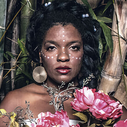 Headshot of Renee Benson, a Black woman with dark brown skin standing in a bamboo forest. The photo only captures her holding pink peonies on her right side with her left shoulder bare and holding up the adornment of a circular gold earring in one ear. Her hair is a crown of braids and there is a geometric shape from her forehead to her nose that continues out to either side of her face made of white paint.