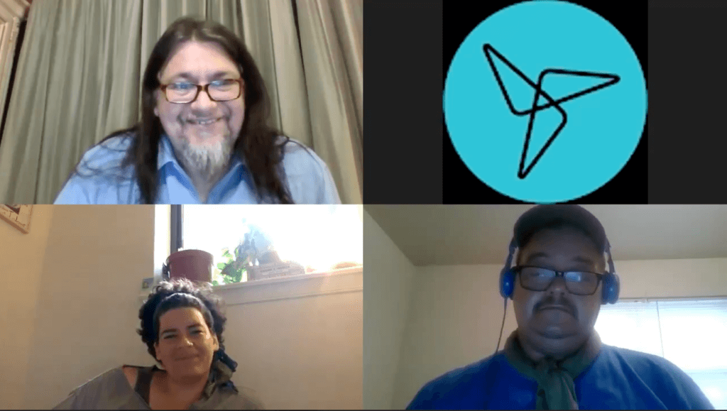 A grid of four screens during a video conference. The upper left quadrant shows a man with long brown hair, glasses, and a medium-length white beard. The upper right quadrant is empty but for an unidentified logo on a turquoise circle. The bottom left quadrant shows a woman with dark hair pulled back with a headband. The bottom right quadrant shows a man with a mustache and glasses wearing a cap and blue headphones.