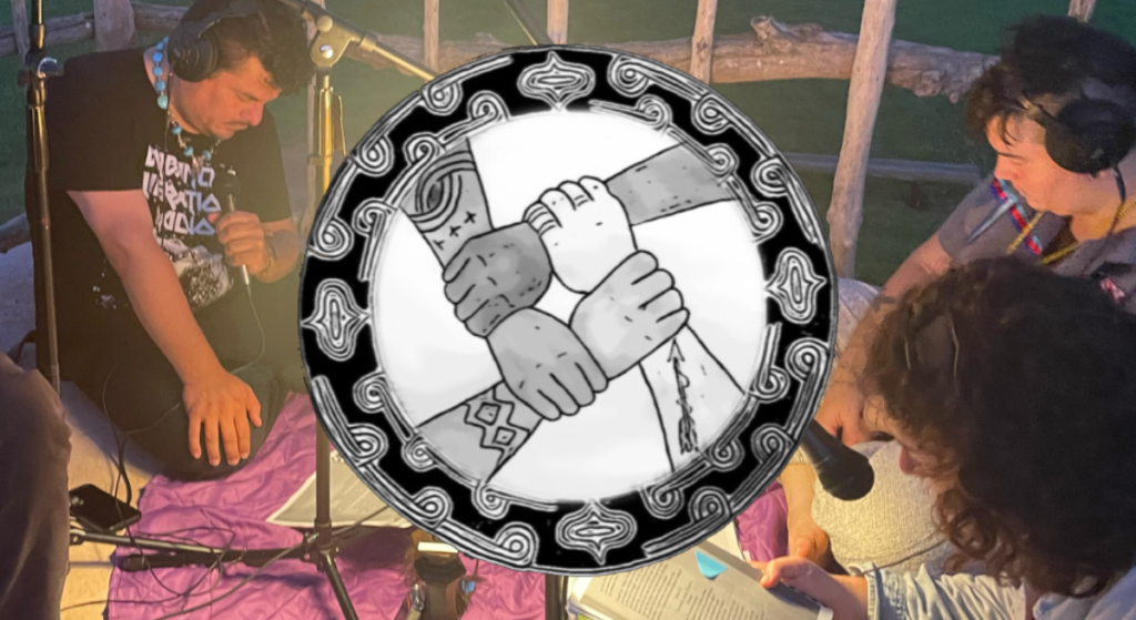 Three actors sit on a blanket in Nanih Bvlbancha at dawn and perform a dramatic reading of the original script of La Fête Du Petit Blé Ou L'héroïsme De Poucha-Houmma.There is a lamp between them and they are looking down at their scripts. Behind them is an irregular fence made out of rough-hewn branches and thin logs. Overlaid on the photo is the black and white logo for the media group.