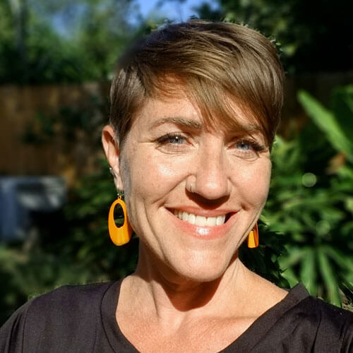 Headshot of Caitlin Strokosch, a light-skinned woman with short brown hair, smiling at the camera against a blurred outdoor background filled with greenery. She is shown from the shoulders up, wearing bright orange earrings and a dark top, with sunlight highlighting her face, giving a warm, natural look.