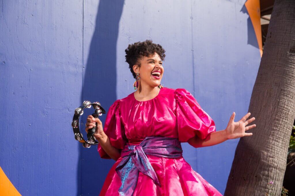 Caramel skinned Black American woman with short curly hair in a hot pink dress, holding a tambourine. Outside; blue building in the background.