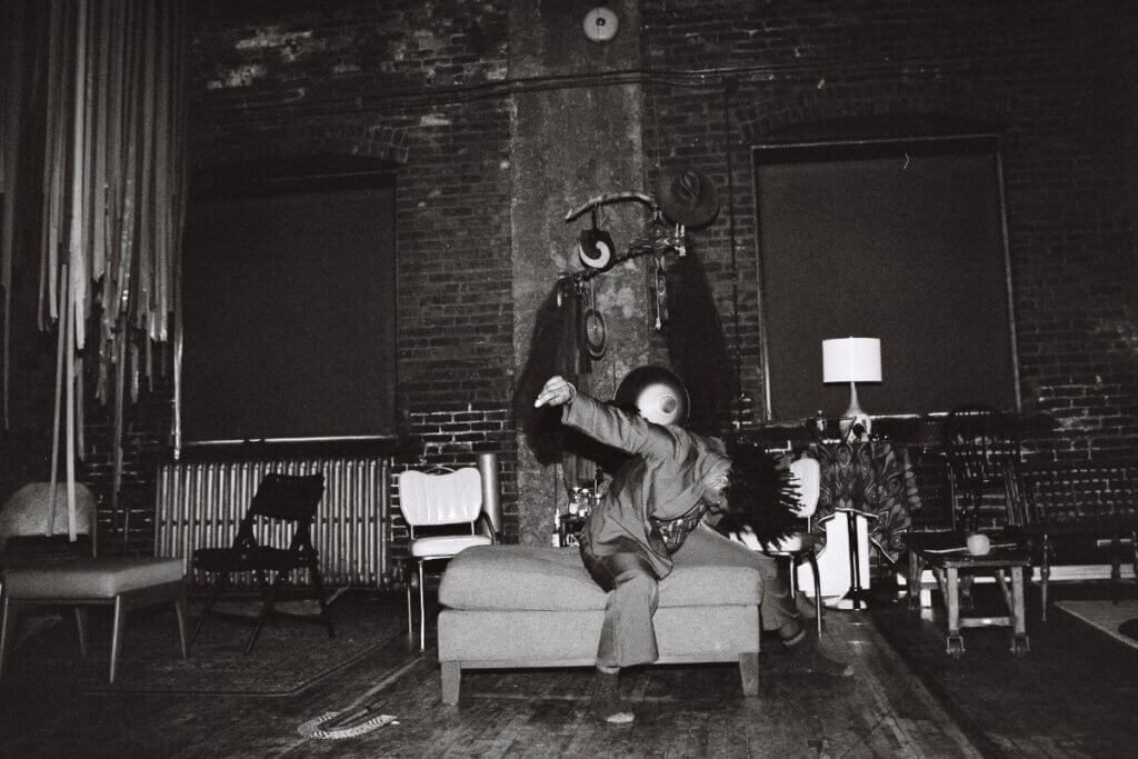 A Black woman is performing a dynamic dance move while seated on an ottoman chaise surrounded by a set depicting a domestic location. She is dressed in a suit and is throwing her head forward, causing her hair to fan out and up. Her arms are spread out behind her as she snaps her fingers.