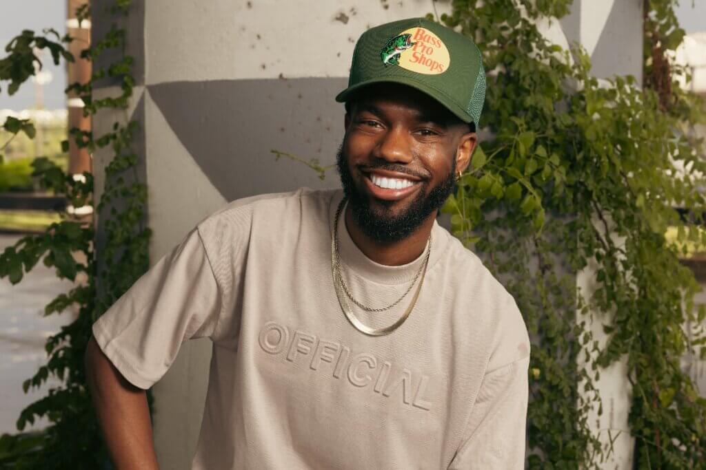 A dark-skinned man sits smiling beneath a concrete overpass covered in leafy green vines. He wears a forest green mesh baseball cap, a beige T-shirt with the word, and light blue ripped jeans. He accessorizes with a gold chain, beaded bracelet, gold rings, and a smartwatch with a brown strap. One arm rests casually on his leg while the other holds the edge of the stool he is sitting on.