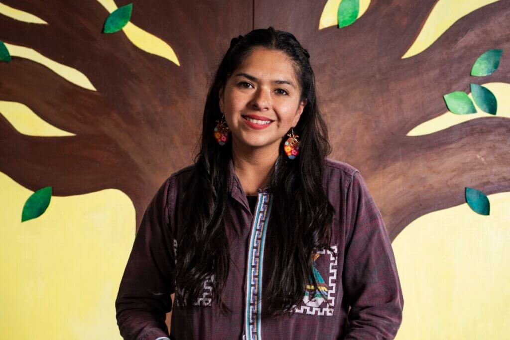 A Mexicana identifying individual with long brown hair wears dangling earrings and a brown button-up shirt with patterned trim and pockets. She stands against a wall mural of a brown tree with green leaves, against a yellow background.