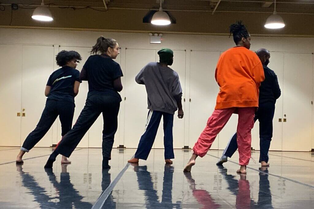 Five dancers in mostly dark clothing (excepting one in bright orange) of mixed genders with their backs to the camera looking over their shoulders to their right in a rehearsal studio with pooled overhead lighting.
