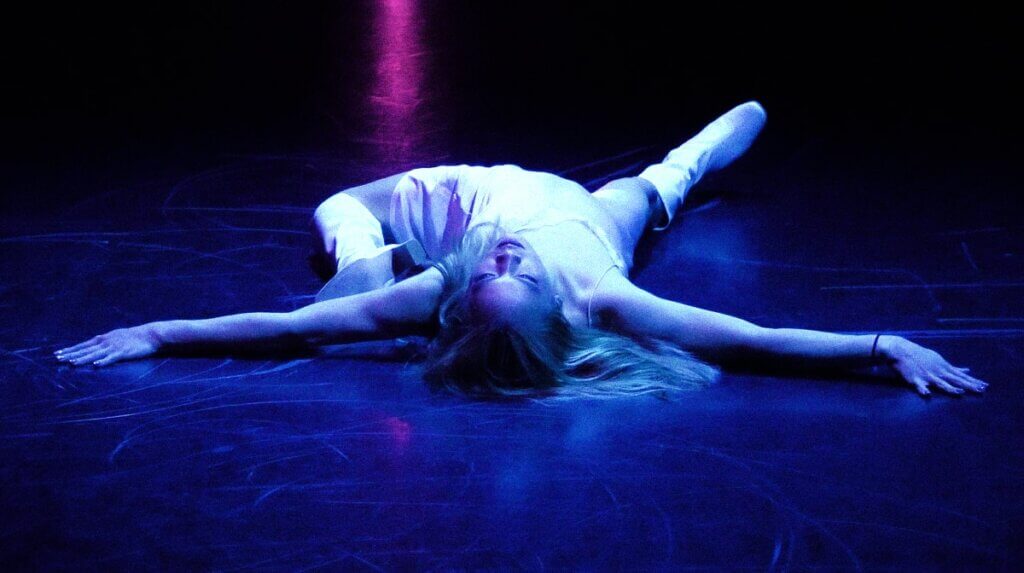 A medium-skinned trans woman with long, bleach-blonde hair is seen lying across a black marley floor in a "death drop." She wears a pink satin slip dress and a pair of white, knee-high gogo boots. Her hair cascades into the floor in the foreground, while her feet trail into the distance. Her arms are spread flat into the ground in full wingspan. Her gaze is set towards the ceiling and she has an easeful look on her face. The main colors that pop out from the photo are shades of pink, blue, and white.