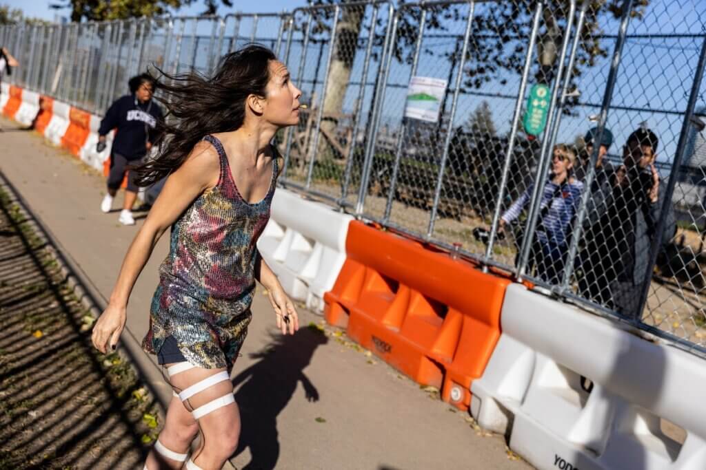 A Yup’ik woman with long dark hair wearing a colorful, sleeveless patterned dress walks purposefully along a pathway lined with orange-and-white barricades. Her legs are wrapped with four strips of white fabric tape, and her hair and dress are blown in the breeze as she looks upward with focus. Behind a chain-link fence, onlookers watch from inside a city park on a sunny day, with shadows and trees visible.