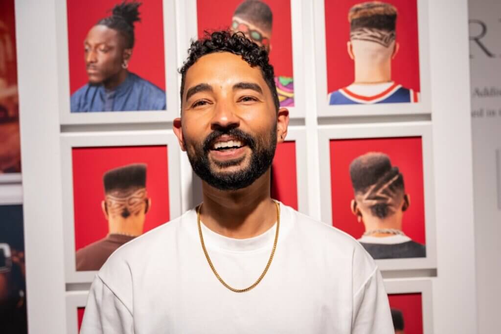 Photo of Gabriel Cortez smiling in front of a photo gallery of Black men's haircuts at the Museum of the African Diaspora (MoAD) in San Francisco. Gabriel is seen wearing a white t-shirt and gold necklace.