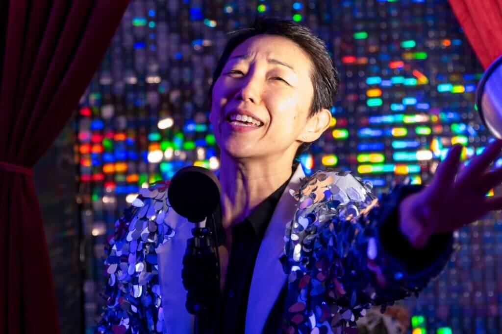 A medium-light skinned Asian woman with short black hair, emotively singing into a stand with a round light at the top. One hand is clutching the light stand, the other hand extends out reaching toward the audience. She is wearing a black button up shirt under an oversized glittery overcoat. The wall behind her is covered with shiny, glittery tiles that are framed by red velvet curtains.