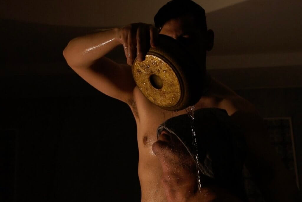 An olive skinned person with black short hair pours water from a flat round copper pan (Baraka bowl) onto the head of another olive skinned person kneeling below. The person receiving the water is shrouded in a light gray thin towel (fouta) folded and placed over their eyes and face. Their skin and the bowl reflect the light, while the rest of the interior space of the dome shaped bathing room (hammam) remains mostly in shadow.