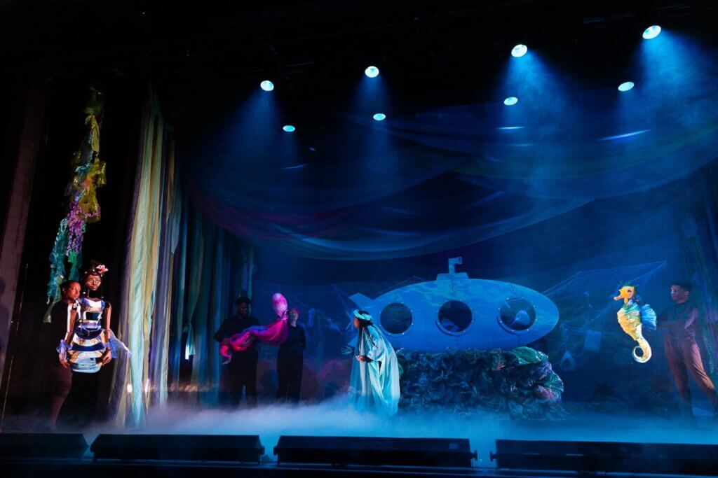 A theatrical underwater scene is depicted on stage, bathed in blue lighting and fog to create an oceanic ambiance. Performers dressed in black operate large, colorful puppets, including black mermaid, a bright pink octopus, and a yellow seahorse. A light blue submarine with round windows and a periscope is set center stage, surrounded by piles of plastic-like debris. Strips of fabric resembling seaweed hang from above and the sides, adding to the immersive, underwater environment.