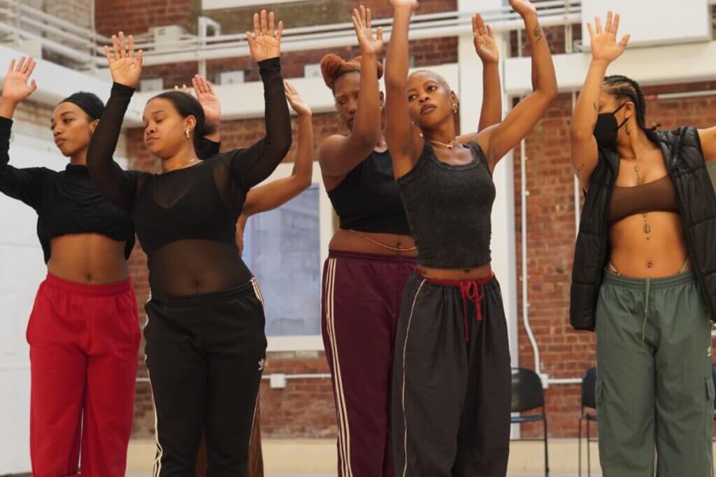 A group of Black femmes in dance clothing stand in a staggered formation with their arms lifted up parallel and pressed out to the sides. They look over their right shoulder.
