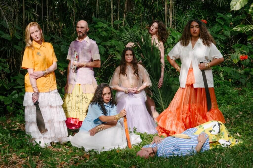 Seven dancers of varying skin colors are dressed in guayaberas (traditional Cuban male dress shirts) and long voluminous skirts. They are sitting, squatting, and laying on the ground in a tropical setting filled with palm fronds. Some are holding machetes and looking into the camera with various expressions of confrontation, surrender and reverence. One dancer's body is totally immersed in palm fronds and only her face is visible.
