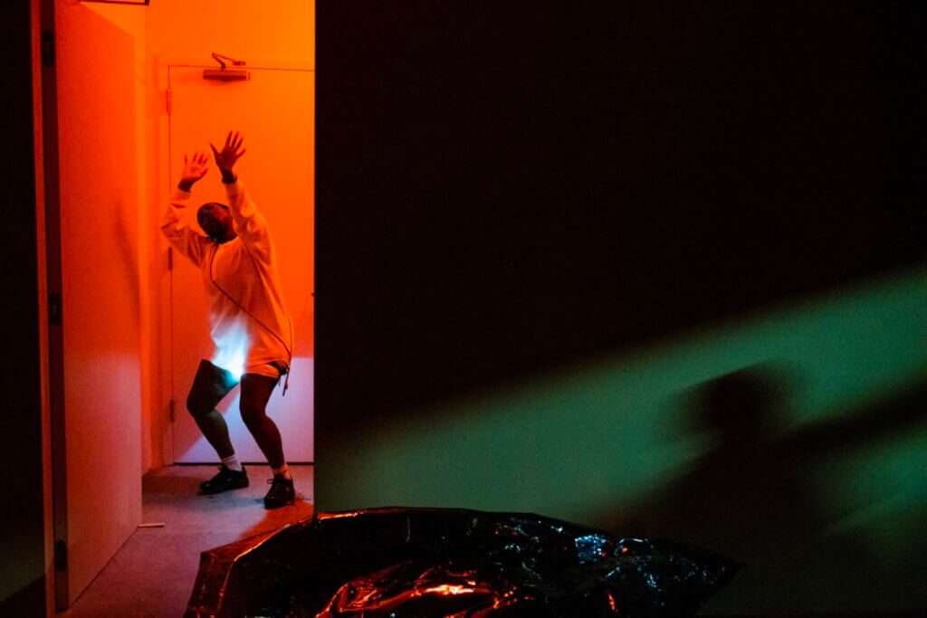 Zinzi Minott standing in an entryway in the left side of the picture with the door ajar dancing in red light, the photographer is in a room on the other side of the doorway that room is dark with green light streaming in from the right a shadow of a person is cast onto the wall a bag made out of double sided mirror multicolor film lies on the floor in the foremost front.