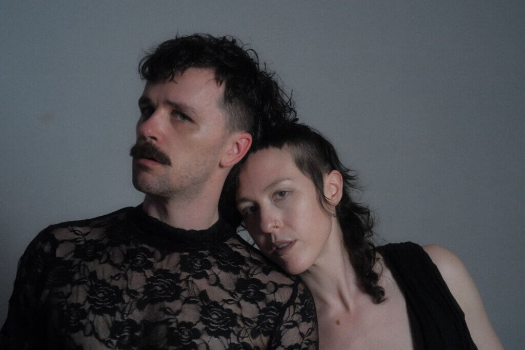 Two white non-binary folks with dark mullets lean their heads in towards the center for a moody headshot with a grey background. Taylor, on the left with a mustache and semi-transparent black lace top, tilts their head to their right with their eyes slanted towards the camera. Anna, in a black low cut black top with a brown birthmark on their chest, leans their chin on taylor’s shoulder and angles their head in towards taylor while pointing their eyes towards the camera.