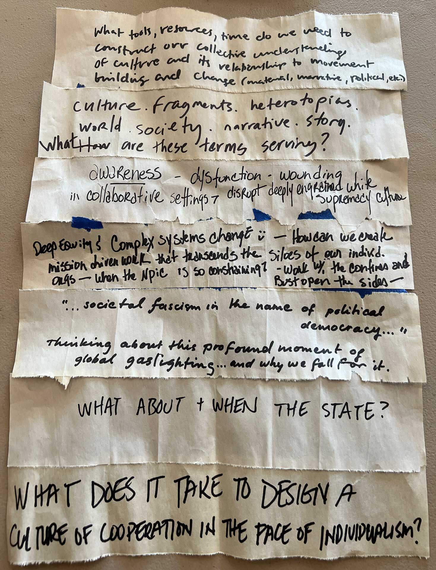 Torn strips of paper laid out in overlapping rows, each handwritten with questions and notes about culture, collective understanding, systems change, fascism, the state, cooperation, and individualism. The separate pieces suggest the ideas can be rearranged and recombined.