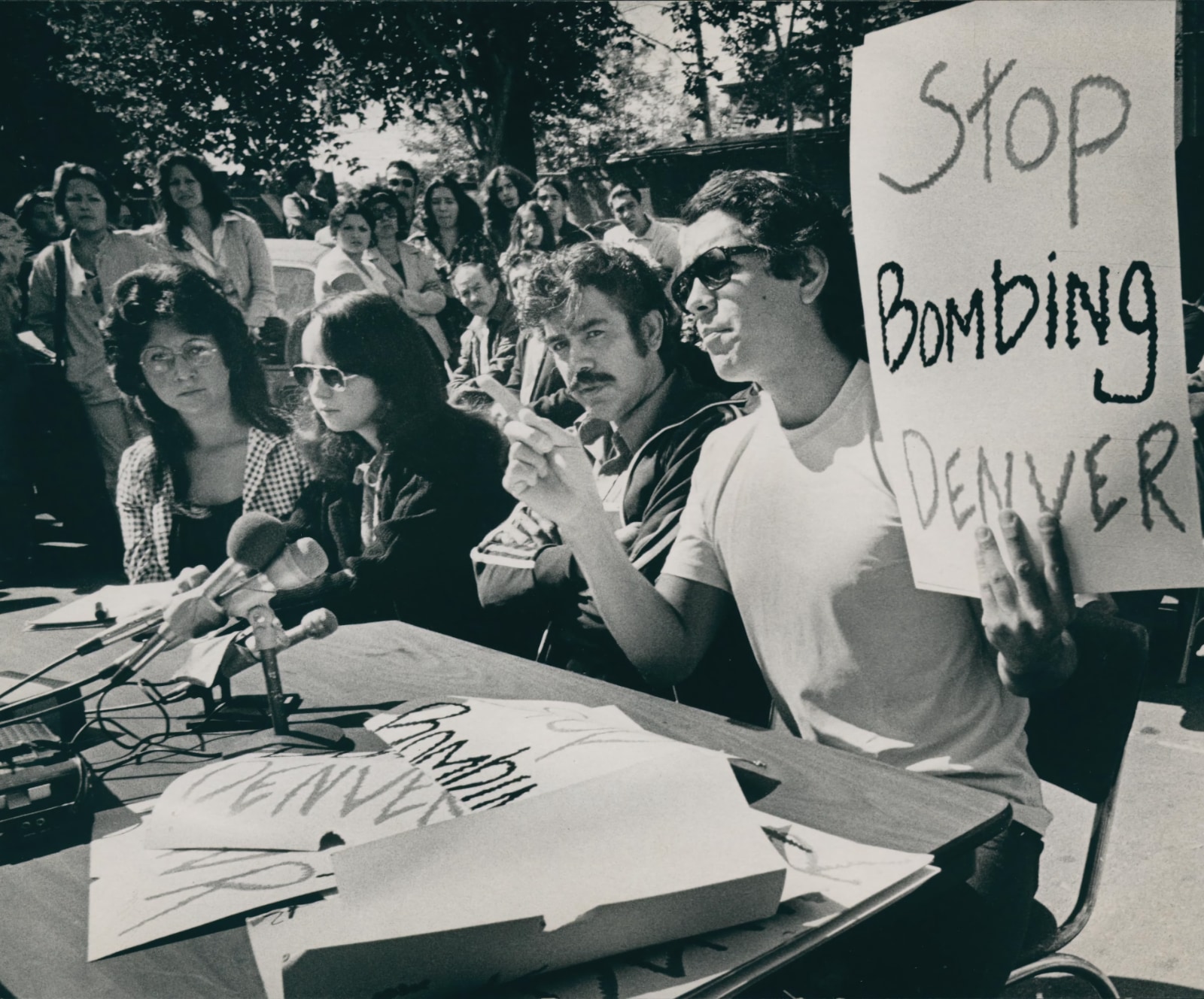 A black and white photo shows four people — two women with long dark hair and two men with dark hair, with one sporting a mustache — sitting in a row behind a small table outdoors. A group of press microphones is assembled in the center of the table. The man sitting on the right side of the row of four is holding up a hand lettered sign with his left hand that reads, "Stop Bombing Denver." He is holding up one finger with his right hand as if making a specific point. Other similarly hand-lettered signs spread in a loose pile on the table in front of him. The other three attendees sit silently. Behind them in the near distance is a crowd of onlookers.