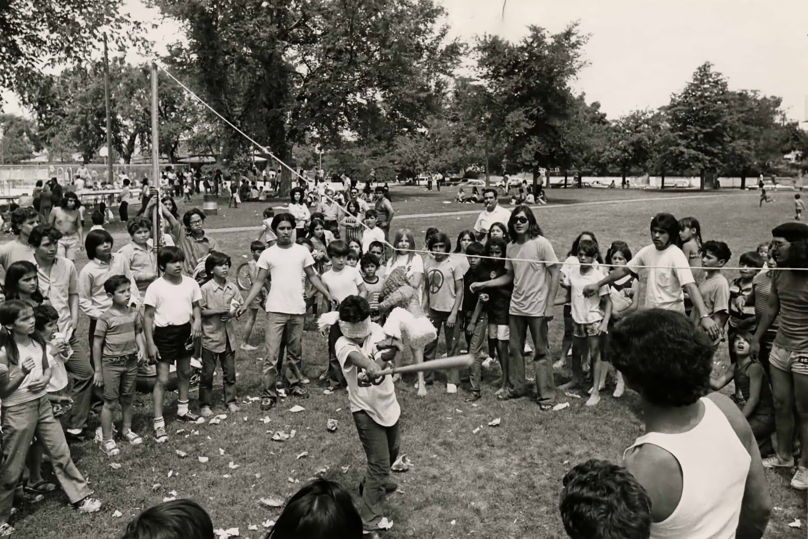 Black and white photograph of a large group of children at a public event in a park. The children are standing in a circle around a hanging piñata, where a small blindfolded boy is swinging a bat in an attempt to hit it. In the background, dozens of people are visiting around tables or seated on blankets spread over the grass.