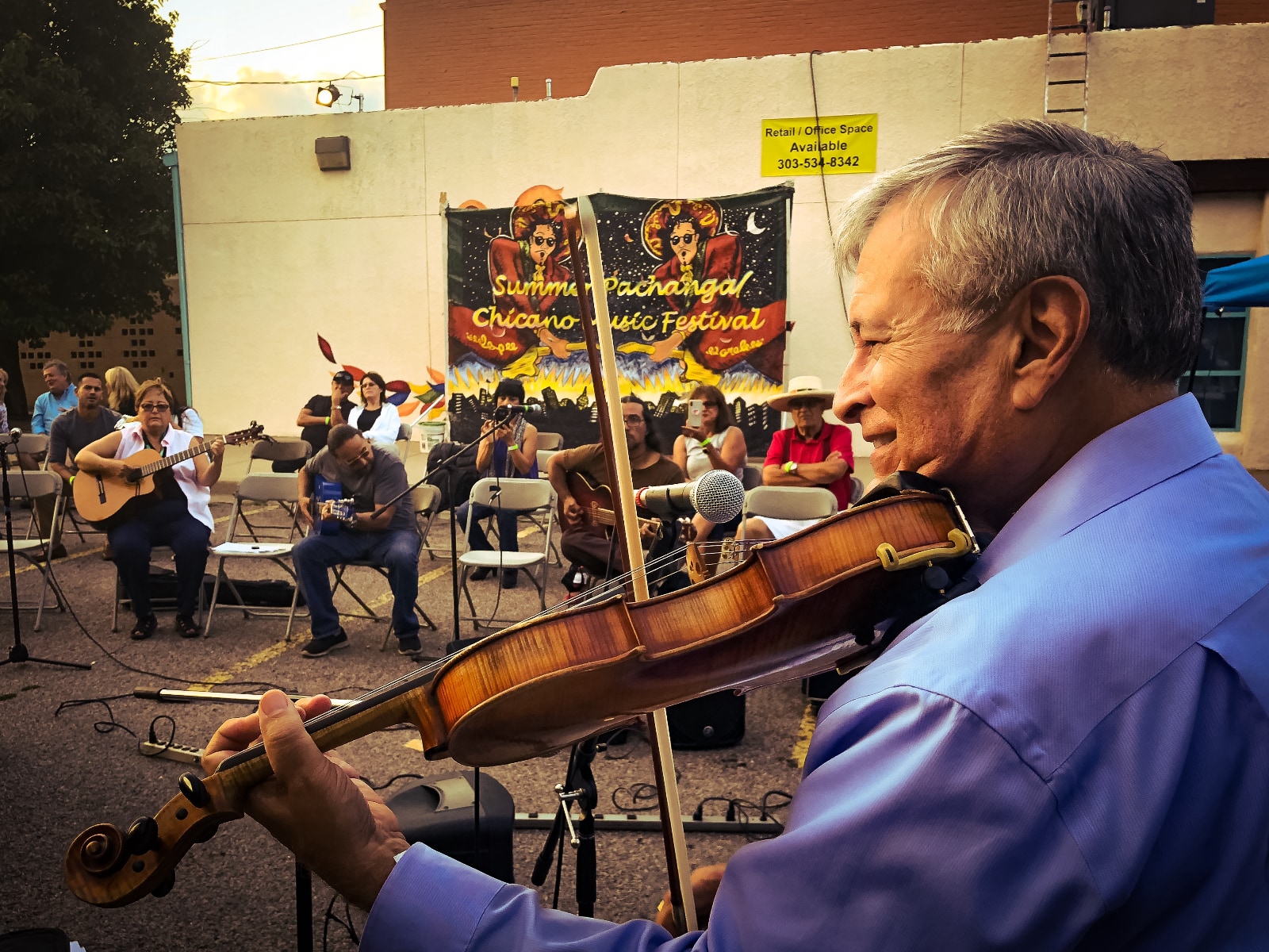 Close up photograph of an older man in profile playing a violin as part of a public performance in a parking lot. He has salt-and-pepper hair and is wearing a blue long-sleeved shirt, and he is smiling as he plays. In the background sitting on folding chairs, are three guitarists accompanying him, and behind them are half a dozen people sitting on folding chairs and listening.