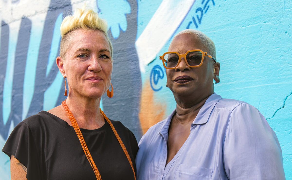 NPN Co-Executive Directors Caitlin Strokosch and Sage Crump stand together outside. The concrete wall behind them is teal and has a mural painted on it, only part of which is visible.