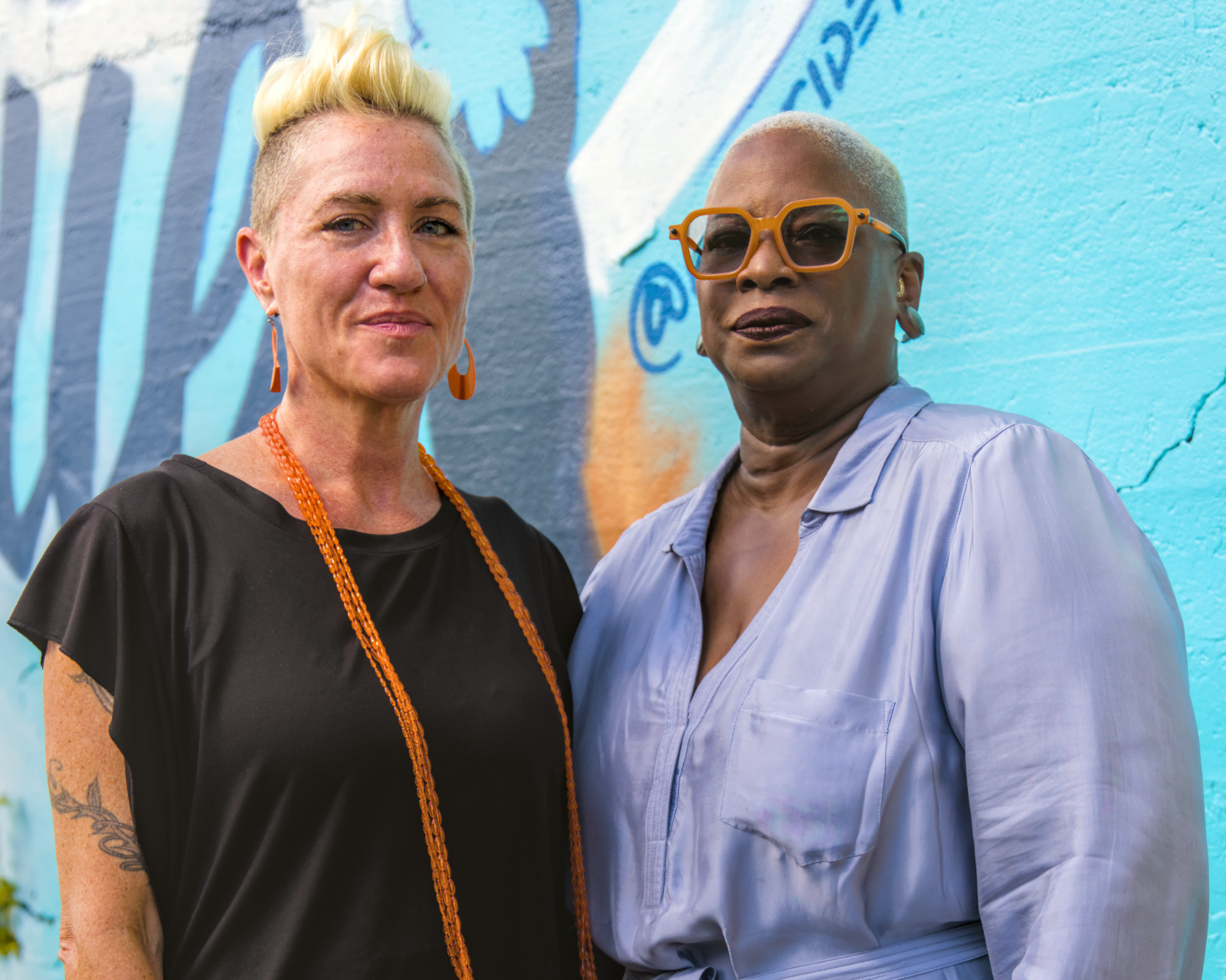 NPN Co-Executive Directors Caitlin Strokosch and Sage Crump stand together outside. The concrete wall behind them is teal and has a mural painted on it, only part of which is visible.