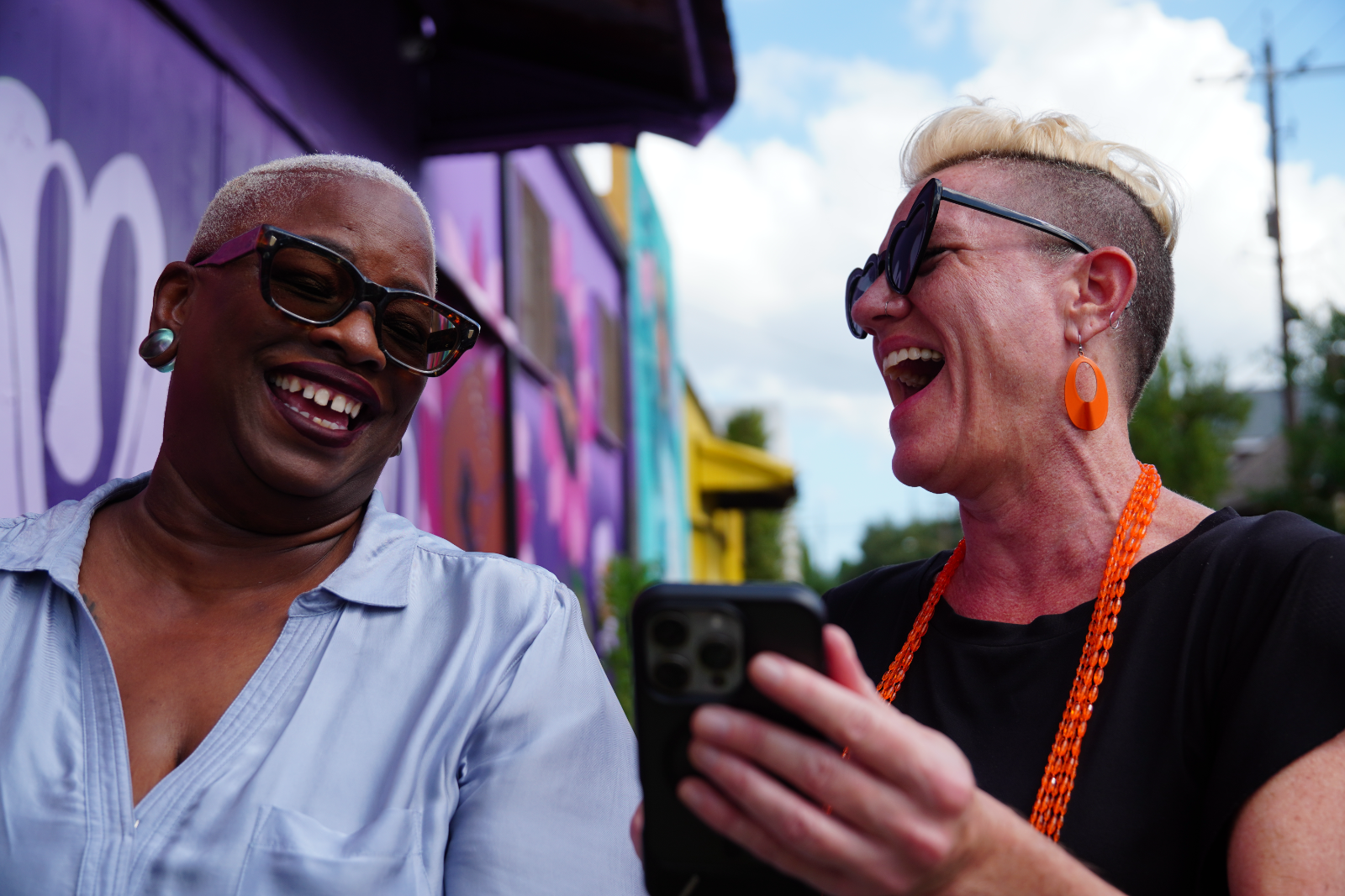 NPN Co-Executive Directors Sage Crump and Caitlin Strokosch stand together outside under a blue sky with large white clouds. They are both laughing at something on Caitlin's phone. Extending into the distance behind them is the wall of a long building, with sections painted in murals of predominantly purple, teal, or yellow.