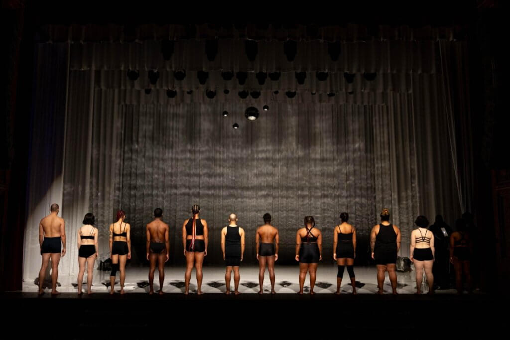Twelve dancers stand in a line on a dimly lit stage, facing away from the audience. Dressed in minimal black attire, they embody unity and vulnerability. A textured curtain and hanging disco balls create atmospheric depth, while subtle lighting enhances their presence. Their diverse body types and hairstyles. In the background, two other cast members add a behind-the-scenes element.