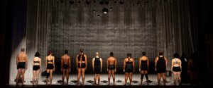 Twelve dancers stand in a line on a dimly lit stage, facing away from the audience. Dressed in minimal black attire, they embody unity and vulnerability. A textured curtain and hanging disco balls create atmospheric depth, while subtle lighting enhances their presence. Their diverse body types and hairstyles. In the background, two other cast members add a behind-the-scenes element.