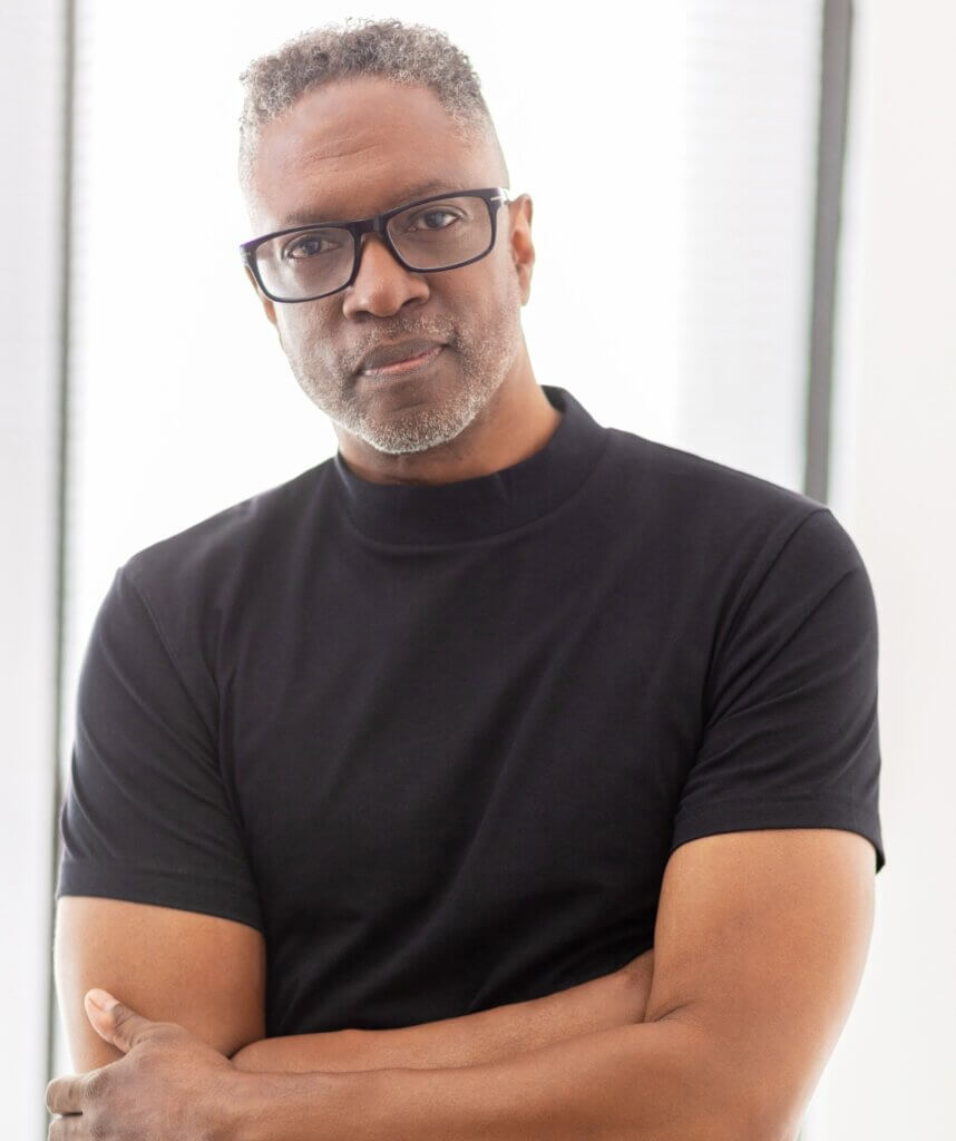 A dark skinned African American man with short naturally curly mixed black and grey hair, wearing a black shirt black rectangular glasses stands against a white background. His arms are crossed, and he is softly smiling.