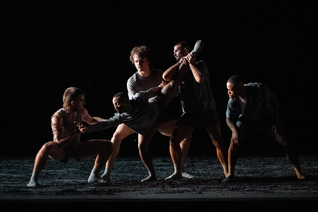 Five dancers on stage in grey costumes, 4 dancers are holding/stretching a central dancer. The scene is dark, there is black material on the floor and the body of the dancers that looks like cinder/ashes.