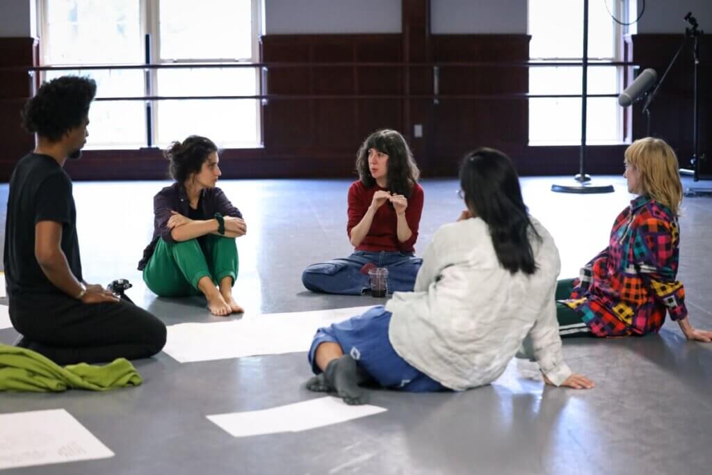 A group of five individuals sit in a circle around a piece of white poster board on a dance studio floor. From Left to right; a Black man kneeling in profile dressed in a black t-shirt and sweatpants, an Arab American woman hugging her knees dressed in bright green sweatpants and a dark purple flannel sits next to him looking over her shoulder at a light-skinned woman speaking -she is the subject of the group’s attention- she is sitting cross legged wearing jeans and a red sweater and using her hands to emphasize her point by bringing her finger tips together, across from her in the circle a Japanese person is sitting with their back to the camera wearing glasses and a white long sleeve shirt and blue sweatpants, next to them and a light-skinned woman is sitting in profile wearing a multi colored flannel jacket. Behind the group a long range microphone is angled at the group on a stand.