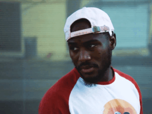 Devonta Ravizee, wearing a white raglan t-shirt with red sleeves and a dirty white baseball cap turned backwards, stands in front of a painted corrugated metal security gate at dusk and stares back over his right shoulder at something off screen.