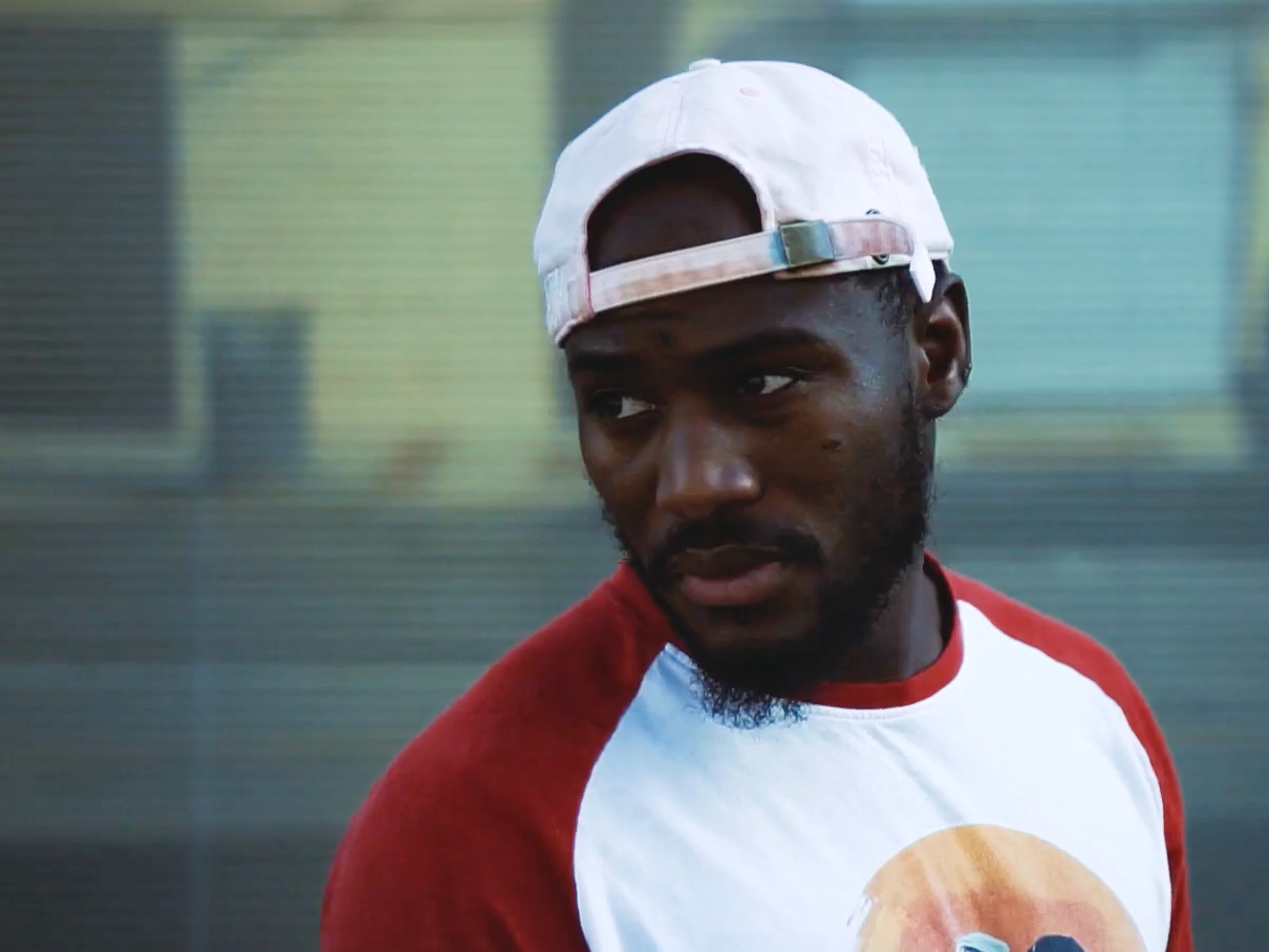 Devonta Ravizee, wearing a white raglan t-shirt with red sleeves and a dirty white baseball cap turned backwards, stands in front of a painted corrugated metal security gate at dusk and stares back over his right shoulder at something off screen.