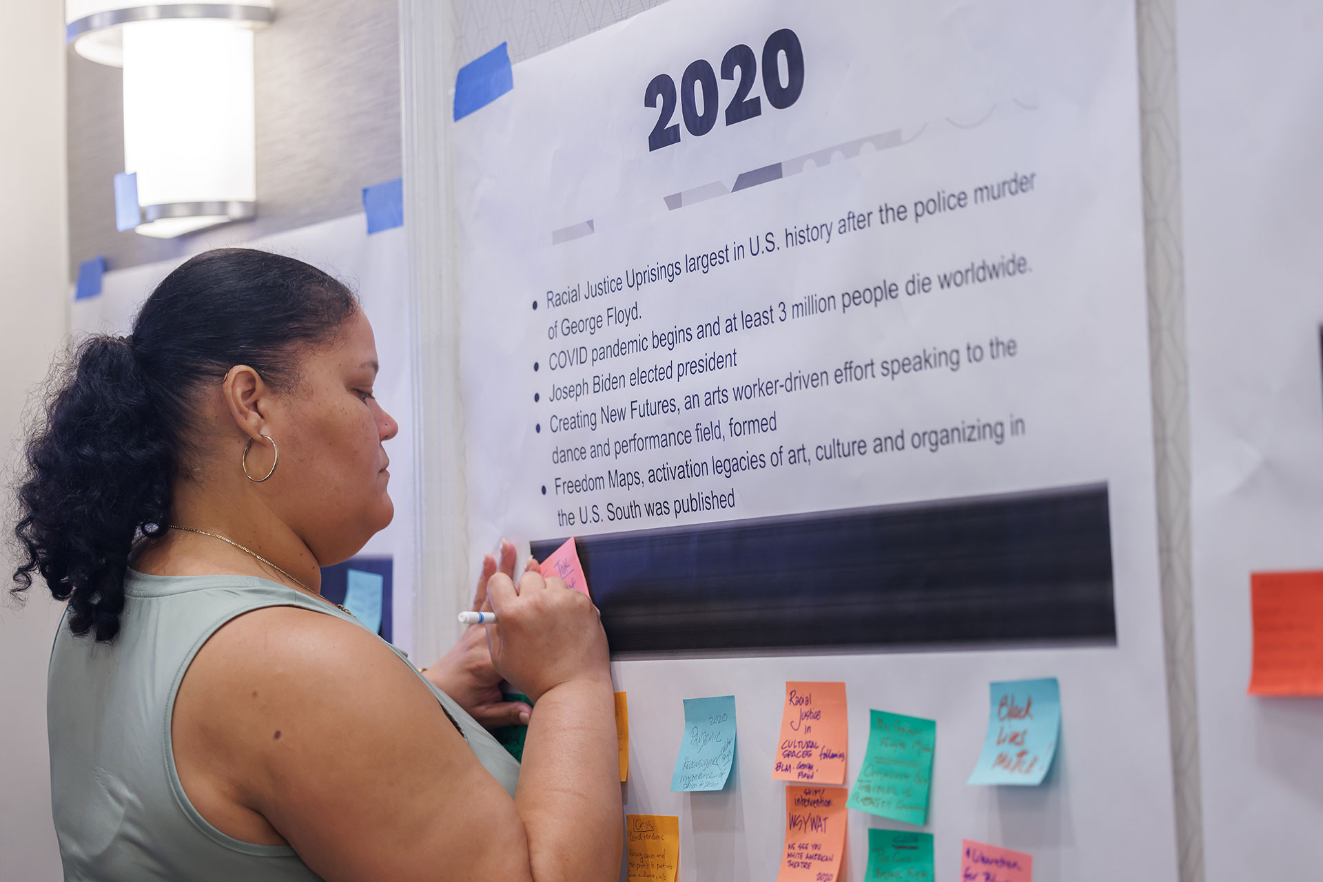 A woman adds a sticky note to a timeline poster titled '2020,' listing events including the racial justice uprisings after George Floyd's murder, the COVID pandemic, and Joseph Biden's election, with colorful sticky notes already placed below.