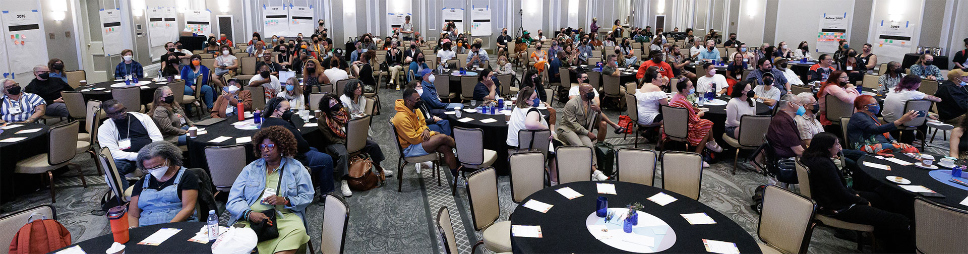 A large ballroom filled with seated attendees, most wearing masks, arranged at round tables facing a timeline display on the walls with years ranging from before 2005 to 2023.