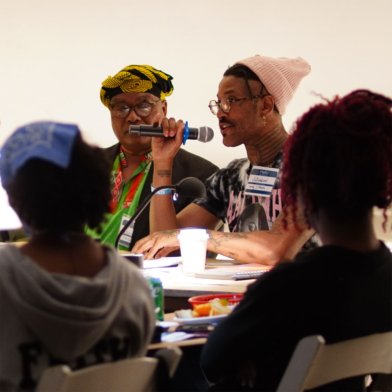 A speaker holds a microphone while addressing the room, with attendees seated at a table in the foreground listening.