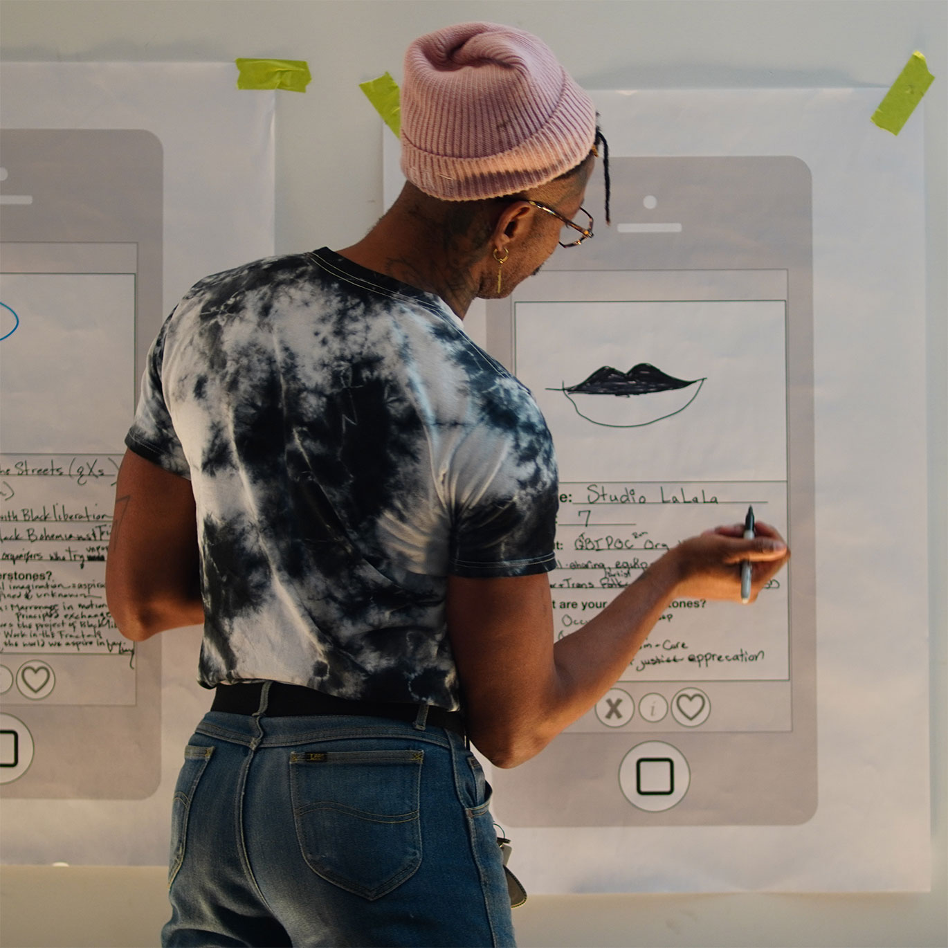 A person in a pink beanie and tie-dye shirt writes on a large paper worksheet shaped like a phone app profile, filling in fields like 'Studio LaLala' and 'QTBIPOC' under a hand-drawn illustration of lips.