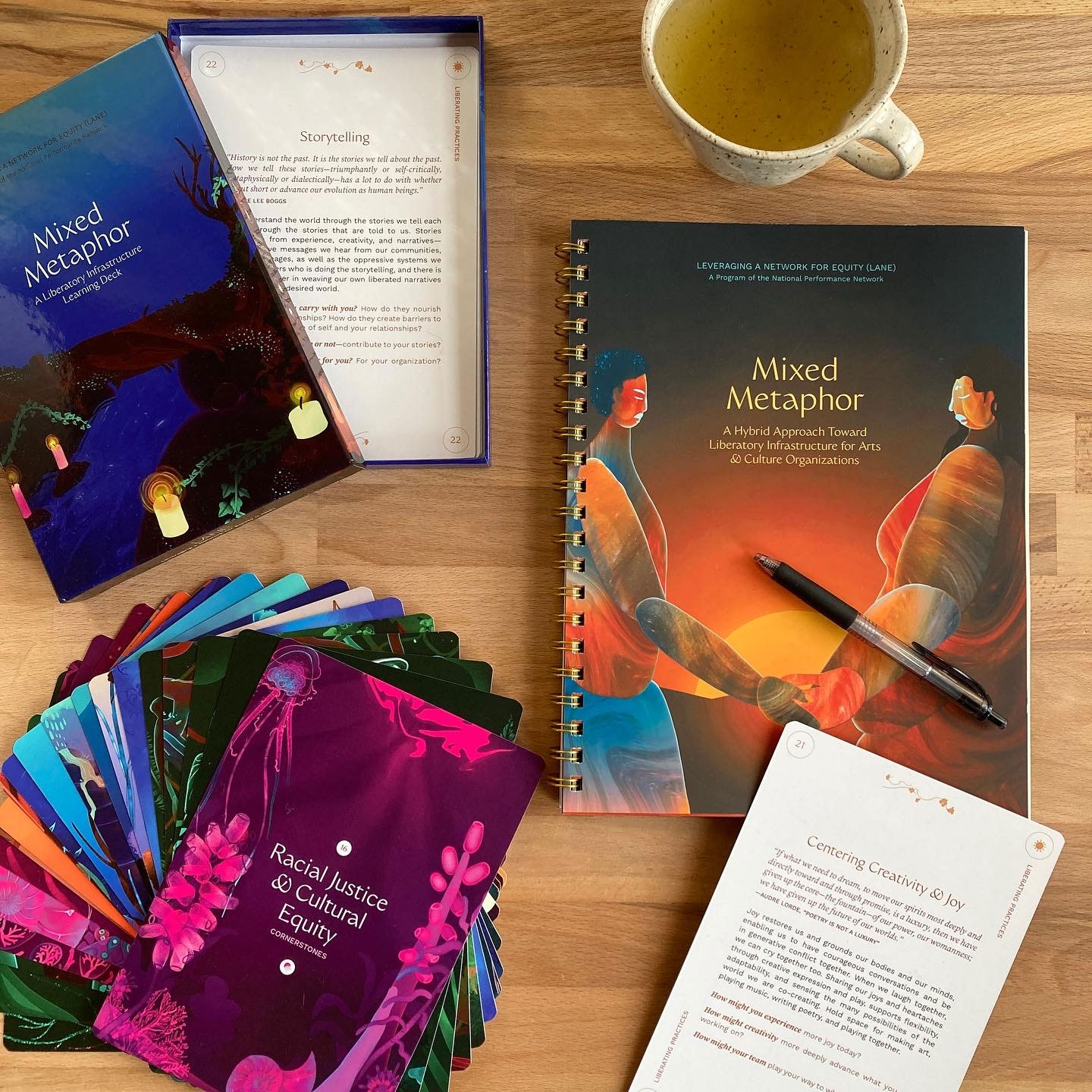 Flat lay of the Mixed Metaphor materials on a wooden table: a spiral-bound guidebook titled 'Mixed Metaphor: A Hybrid Approach Toward Liberatory Infrastructure for Arts & Culture Organizations' with a pen resting on it, a fanned deck of illustrated cards including 'Racial Justice & Cultural Equity,' a card box labeled 'A Liberatory Infrastructure Learning Deck,' an open card titled 'Storytelling,' another card titled 'Centering Creativity & Joy,' and a cup of tea.