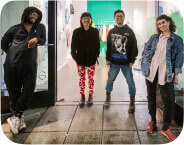 A group of four people outside a gallery with large windows. From left to right: a Black person in a black jumpsuit, an Asian person in red and white pants and black blouse, an Asian person in jeans and graphic hoodie, and a white person in black jeans, pink and white striped blouse and denim jacket.